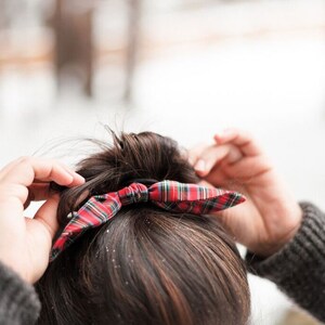 May include: A red, green, and white plaid bow hair accessory is being adjusted. The bow is tied on top of dark brown hair. The background is blurred, with a snowy, wintery scene.