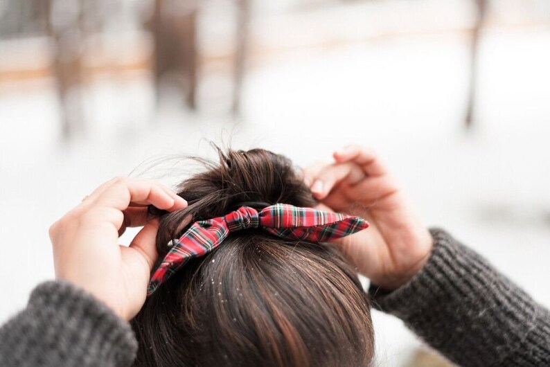May include: A red, green, and white plaid bow hair accessory is being adjusted. The bow is tied on top of dark brown hair. The background is blurred, with a snowy, wintery scene.