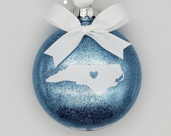 May include: A blue glitter Christmas ornament with a white outline of the state of North Carolina and a small heart in the center. The ornament is hanging from a white ribbon.