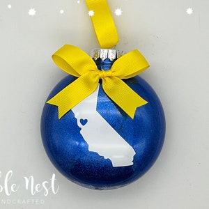 May include: Blue Christmas ornament with a white outline of the state of California and a small heart inside. The ornament has a yellow bow and a yellow string for hanging.