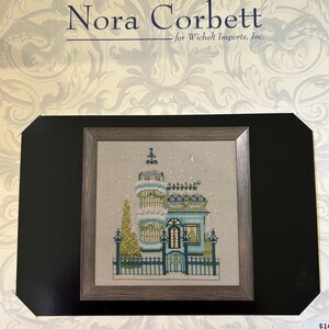 May include: A framed cross-stitch design of a Victorian house with a light blue facade, a green roof, and a Christmas tree. The design is by Nora Corbett. The frame is a light brown color.