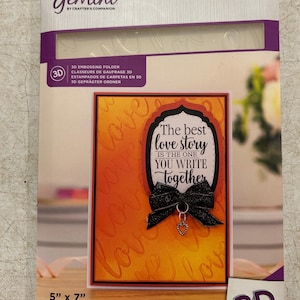 May include: A Gemini 3D embossing folder package. The package features a card with an orange background and the word "love" embossed on it. The card has a black and white oval with the text "The best love story is the one you write together". The card measures 5" x 7".