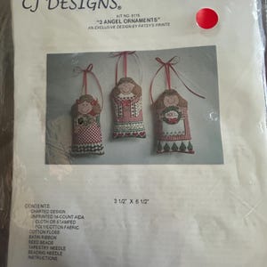 May include: A counted cross stitch kit by CJ Designs, featuring three angel ornaments. The kit includes charted designs, fabric, floss, ribbon, beads, and needles. The package dimensions are 3.5 inches by 6.5 inches.
