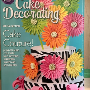 May include: A cake decorating book titled "Cake Decorating" by Wilton. The cover features a two-tiered cake with a zebra print design, adorned with colorful, flower-shaped decorations. The book's special section is "Cake Couture!"