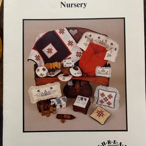 May include: A cross-stitch pattern booklet titled "American Country Nursery" featuring a variety of cross-stitch designs, including a quilt, pillows, and a basket. The booklet is published by T.H.R.E.A.D Connections and has the product number MJ 831.