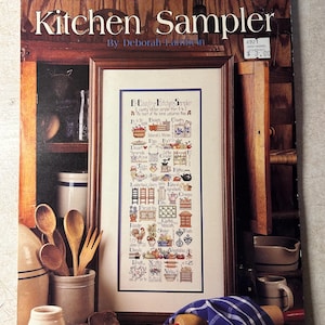May include: A vintage "Kitchen Sampler" cross-stitch pattern booklet by Deborah, featuring a framed sampler with kitchen-themed motifs. The cover displays the title, author, and price, with a rustic wooden background and kitchen utensils.