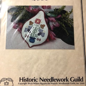 May include: A white fabric ornament with a floral design in shades of blue, red, and green. The ornament is shaped like a pear and has a small blue pot at the bottom. The text "Sampler Ornament 2000" is at the top of the image. The text "Historic Needlework Guild" is at the bottom of the image.