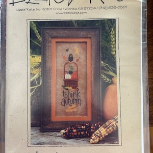 May include: Cross stitch pattern featuring a birdhouse with a bird perched on top, surrounded by leaves and a basket of fruit. The text "think autumn" is below the birdhouse. The pattern is framed and displayed on a wooden surface with corn cobs.