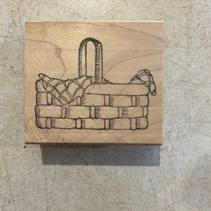 May include: A wooden stamp featuring a detailed illustration of a woven basket with a handle. The basket contains a checkered cloth. The stamp is made of light-colored wood and is ideal for crafting and scrapbooking projects.