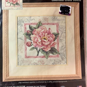 May include: A Dimensions counted cross stitch kit titled Peony Blossom, featuring a pink peony flower design on ivory Aida fabric. The packaging displays the finished 10 inch by 10 inch artwork, including cotton thread, a needle, and instructions.