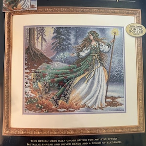 May include: A framed cross-stitch kit titled "Woodland Enchantress" by Ruth Sanderson. The artwork depicts a woman in a flowing green and white gown, holding a staff, in a snowy forest scene. The kit measures 14 x 12 inches.