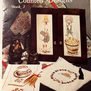 May include: Book cover for "Jo Sonja's Counted Designs, Book 3". The cover features a vintage-style illustration of two geese, one wearing a hat and the other a bonnet, with a quilt pattern in the background.