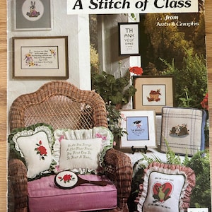May include: A book titled "A Stitch of Class" from Arts & Graphs, featuring cross-stitch patterns. The book cover shows a wicker chair with pillows featuring cross-stitch designs, including a bunny, a frog, and a graduation cap.