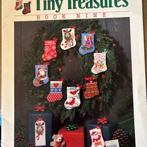 May include: A book titled "Tiny Treasures Book Nine" by Linda Gillum featuring cross stitch patterns for Christmas stockings. The cover shows a wreath of pine branches with nine different stockings decorated with various Christmas themes, including a moose, an angel, a Santa Claus, a cardinal, and a plaid design. The book is published by Dimensions.