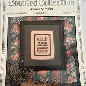 May include: A cross-stitch pattern booklet titled Pat Rogers' Counted Collection Anna's Sampler. The cover features a framed sampler with the year 1992, the letters ABCD, and the initials PR, set against a floral wallpaper background with glasses.