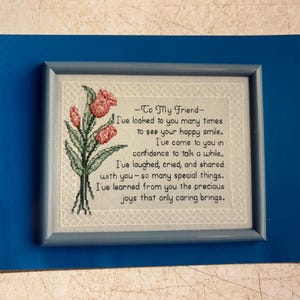 May include: A framed cross-stitch with a blue frame and a white background. The cross-stitch features a floral design with pink flowers and green leaves. The text reads: "To My Friend - I've looked to you many times to see your happy smile. I've come to you in confidence to talk a while. I've laughed, cried, and shared with you - so many special things. I've learned from you the precious joys that only caring brings."