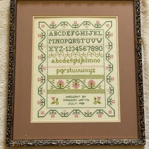 May include: A framed cross-stitch sampler with the alphabet, numbers, and the text "Wrought by Margery Watts July 1981". The sampler is surrounded by a decorative frame with a gold finish.