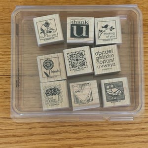 May include: A clear plastic container holding nine wooden stamps. The stamps feature various designs, including floral patterns, a bird on a branch, an owl, and the words "for you," "thank U," "thinking of you," "friends," "eat cake," and "love."