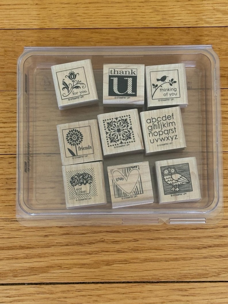 May include: A clear plastic container holding nine wooden stamps. The stamps feature various designs, including floral patterns, a bird on a branch, an owl, and the words "for you," "thank U," "thinking of you," "friends," "eat cake," and "love."