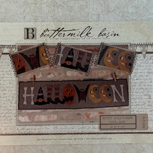 May include: A rectangular Batty Halloween wool mat pattern package by Buttermilk Basin. The design features a brick wall background with a banner spelling HALLOWEEN using pumpkins, moons, bats, and witch hats. It measures 39.5 inches by 10 inches.