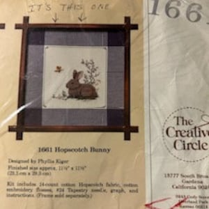 May include: A cross-stitch kit featuring a brown bunny in a garden setting, framed within a dark wooden border. The kit includes fabric, thread, and instructions. The finished size is approximately 11.25" x 11.25". The text "IT'S THIS ONE" is above the image.
