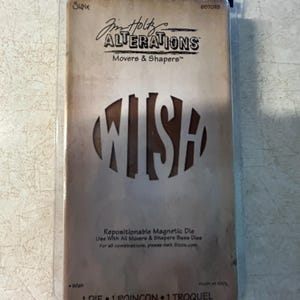 May include: A Sizzix Tim Holtz Alterations Movers & Shapers repositionable magnetic die in original clear plastic packaging. The tan card insert features the word WISH cut out in a circular center, with branding text and product details printed.