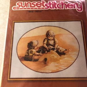 May include: A Sunset Stitchery needlepoint kit titled Sand Castle in a clear plastic bag. The packaging features an oval illustration of two children playing in the sand with buckets. The text reads sunset stitchery and SAND CASTLE.