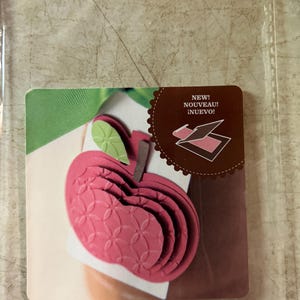 May include: A red apple-shaped card with a green leaf and stem. The card is layered and has a patterned design. The text "NEW! NOUVEAU! INUEVO!" is on a brown circle with a scalloped edge.