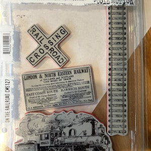 May include: A set of four vintage-style rubber stamps featuring a train, railroad tracks, and a railroad crossing sign. The stamps are packaged in a clear plastic case with the text "On the Railroad CMS127" and "Stampers Anonymous" printed on the front. The stamps are part of the "Jim Holtz Collection".