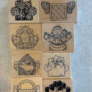 May include: A collection of eight wooden rubber stamps with various designs. The stamps feature images of a snowflake basket, a butterfly and lattice design, an Easter egg, a snowman, a pumpkin with leaves, a pumpkin in a basket, a woven basket, and balloons.