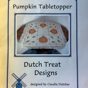 May include: A white cross-stitch table topper with a pumpkin design. The table topper is surrounded by a brown wooden table. The text "Pumpkin Tabletopper" is at the top of the image. The text "Dutch Treat Designs" is in the center of the image. The text "designed by Claudia Dutcher" is at the bottom of the image.