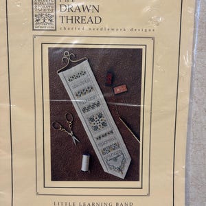 May include: A cross-stitch pattern kit titled "The Acorn Sampler" by The Drawn Thread. The kit includes a needlework design with the alphabet, scissors, thread, and other tools. The design is on a cream-colored background with a brown border.