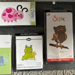 May include: Three die-cutting templates for crafting. The first template features a pink ladybug with black spots and antennae. The second template features a green frog with a flower. The third template features a brown owl with yellow eyes perched on a branch. All three templates are labeled "Sizzlits Die Poinçon Troquel".