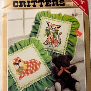 May include: A craft kit featuring two cross-stitch pillows with animal designs, a cat and a dog, surrounded by green ruffled fabric. A small brown teddy bear with a bow is also present. The kit is labeled "Calico Critters" and "Yours Truly Counted Cross Stitch."