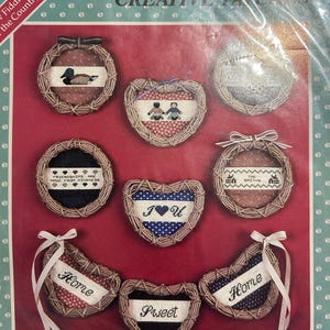 May include: A package of Creative Patch cross-stitch kits featuring various designs. The kits include round and heart-shaped frames made of woven material, with embroidered patterns and text such as "Home," "Sweet," and "I ♥ U." The background is a red surface.