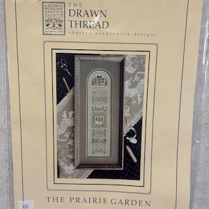 May include: A cross-stitch pattern titled "The Prairie Garden" by The Drawn Thread. The design is framed and features a detailed needlework design with a house, flowers, and other elements. The package includes scissors and a needle.