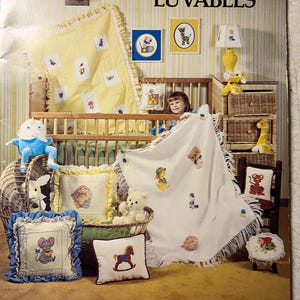 May include: A book titled "Luvables" with a yellow and white striped cover. The book is open to a page showing a nursery with a crib, a rocking chair, and various stuffed animals and pillows. The pillows and stuffed animals are decorated with cross-stitch embroidery. The book is for sale for $4.00.