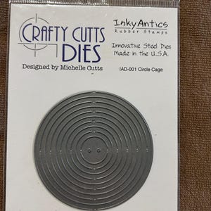 May include: A gray steel die cut in the shape of a circle cage. The packaging reads "Crafty Cutts Dies" and "Innovative Steel Dies Made in the U.S.A." The die is designed by Michelle Cutts and is labeled IAD-001 Circle Cage.