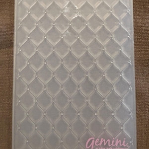 May include: A clear, rectangular embossing folder with a quilted pattern. The design features a grid of diamond shapes with small, raised dots at each intersection. The word "Gemini" is printed in pink script at the bottom, with "by Crafter's Companion" below.
