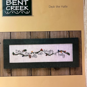 May include: A cross-stitch pattern featuring a green wreath with red berries and white lights. The text "Deck the halls with boughs of holly" is stitched in white thread.