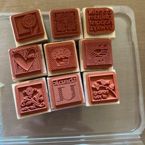 May include: A collection of nine red rubber stamps with wooden blocks. Designs include a sunflower, a heart, an owl, a floral pattern, a cupcake, and the letter "U". Some stamps have text, including the words "love" and "joy to".