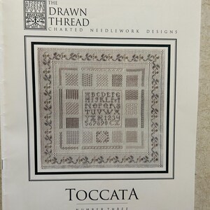 May include: A printed needlework design titled "TOCCATA NUMBER THREE" by The Drawn Thread. The cover features a detailed sampler with various stitch patterns, letters, and numbers. The design is framed with a decorative border.