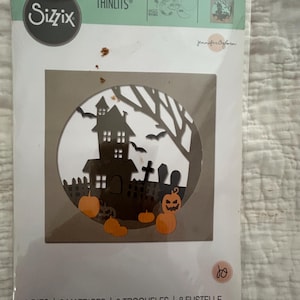 May include: Sizzix Thinlits die set featuring a Halloween-themed design. The die-cut design includes a haunted house, pumpkins, bats, and a graveyard scene. The package includes 8 dies and is designed for crafting projects.