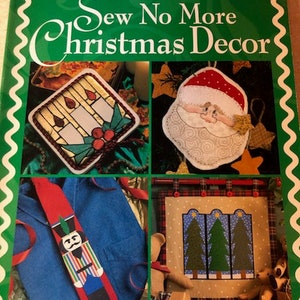May include: A book titled "Sew No More Christmas Decor" by Leisure Arts Presents Wonder-Under. The book cover features four images of Christmas-themed embroidery projects: a candle holder, a Santa Claus, a nutcracker, and a trio of Christmas trees.