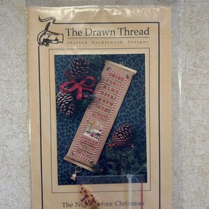 May include: A craft kit for "The Night Before Christmas" cross-stitch design by The Drawn Thread. The kit includes a pattern, beads, and a bell. The image shows a finished cross-stitch design with a Christmas theme, pinecones, and a red bow.