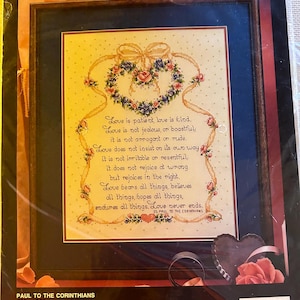 May include: A Bucilla counted cross-stitch kit featuring a framed design with a heart-shaped floral wreath and the text "Love is patient, love is kind." The kit measures 11" x 14" and includes Aida cloth and embroidery floss.