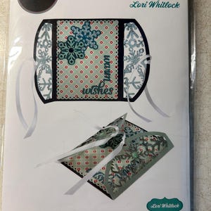 May include: Sizzix Thinlits die set by Lori Whitlock. The package features a die-cut design with snowflakes and the words "warm wishes". The set includes 8 dies for crafting projects. The design is in shades of teal, red, and white.