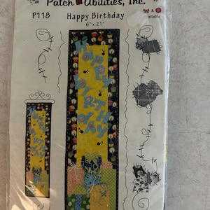 May include: A Patch Abilities, Inc. P118 Happy Birthday sewing pattern in a plastic sleeve. The pattern features a 6 inch by 21 inch vertical banner design with a yellow center, blue lettering, cupcake motifs, and gift boxes on a black background.