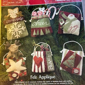 May include: A set of six felt Christmas ornament kits featuring a snowflake, a snowman, a tree, a heart, a star, and a candy cane. The kits include felt, beads, charms, thread, and instructions. The ornaments are approximately 5.5 inches long and can be finished as pockets or stuffed.