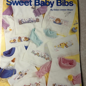 May include: A collection of white baby bibs and shirts with embroidered designs. The bibs feature phrases like "Sweet Baby" and "My Mommy loves me!" along with cartoon characters and illustrations. The cover reads "Sweet Baby Bibs."